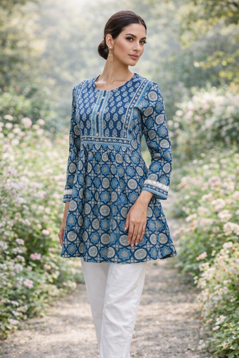 May include: A woman wearing a long-sleeved, blue tunic with a white and gold pattern, paired with white pants. The tunic has a round neckline and a flared hem. The sleeves are long and end with a white lace trim. The woman is standing on a path surrounded by greenery.