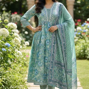 May include: A light green floral print ensemble featuring a long-sleeved tunic, matching pants, and a flowing scarf. The outfit has blue floral accents and intricate detailing around the neckline and hem. The outfit is worn outdoors.