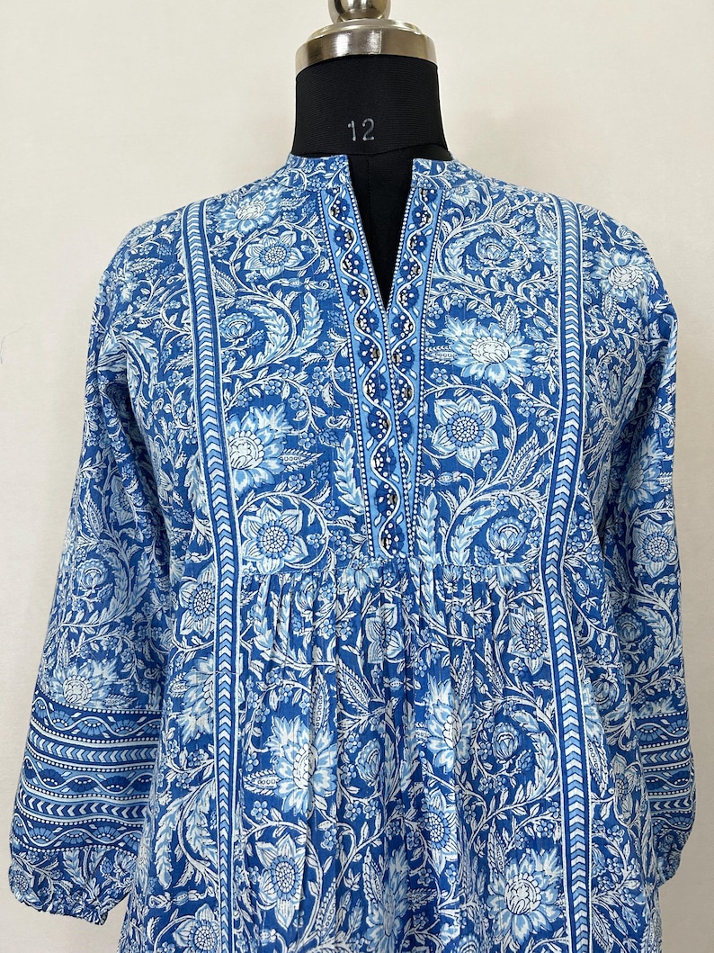 May include: A blue and white floral print long-sleeved tunic with a V-neck and a beaded trim. The tunic has a gathered waist and a relaxed fit.