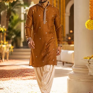 Könnte beinhalten: Ein langärmeliger, knielanger brauner Kurta mit Stehkragen, einer Knopfleiste mit aufwendiger silberner Stickerei und verstreuten kleinen silbernen Blumenmotiven, kombiniert mit einer locker sitzenden, cremefarbenen Dhoti-Hose.