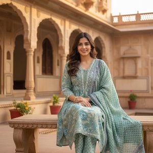 May include: A light green floral print salwar kameez set. The set includes a tunic top, matching pants, and a dupatta. The outfit features a V-neckline and is embellished with silver accents. The model is sitting on a stone bench.