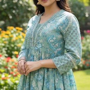 May include: A light teal dress with a floral pattern in shades of blue and white. The dress features a V-neckline, long sleeves, and decorative embroidery along the neckline and cuffs. The dress has a flowing, relaxed fit.