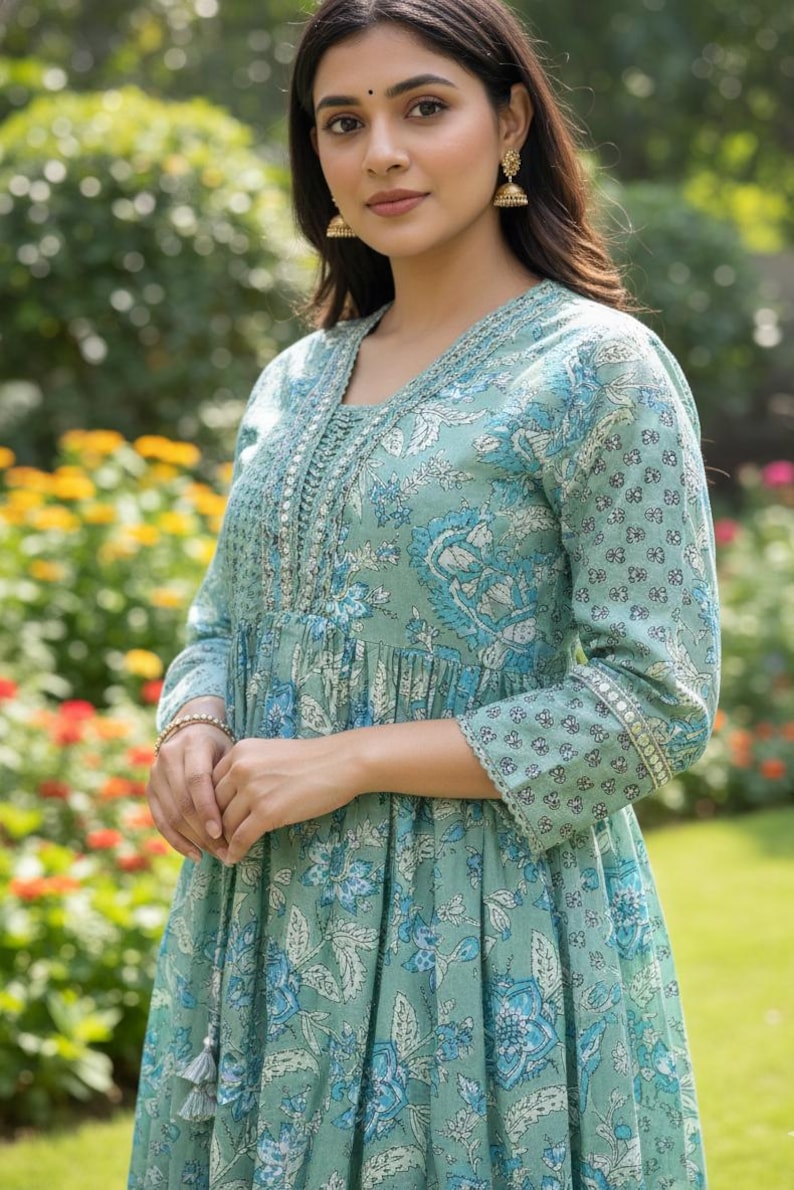 May include: A light teal dress with a floral pattern in shades of blue and white. The dress features a V-neckline, long sleeves, and decorative embroidery along the neckline and cuffs. The dress has a flowing, relaxed fit.