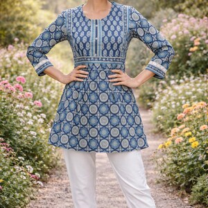 May include: A woman wearing a blue tunic with a geometric pattern and white pants. The tunic has long sleeves and a round neckline. The background is a garden with flowers.