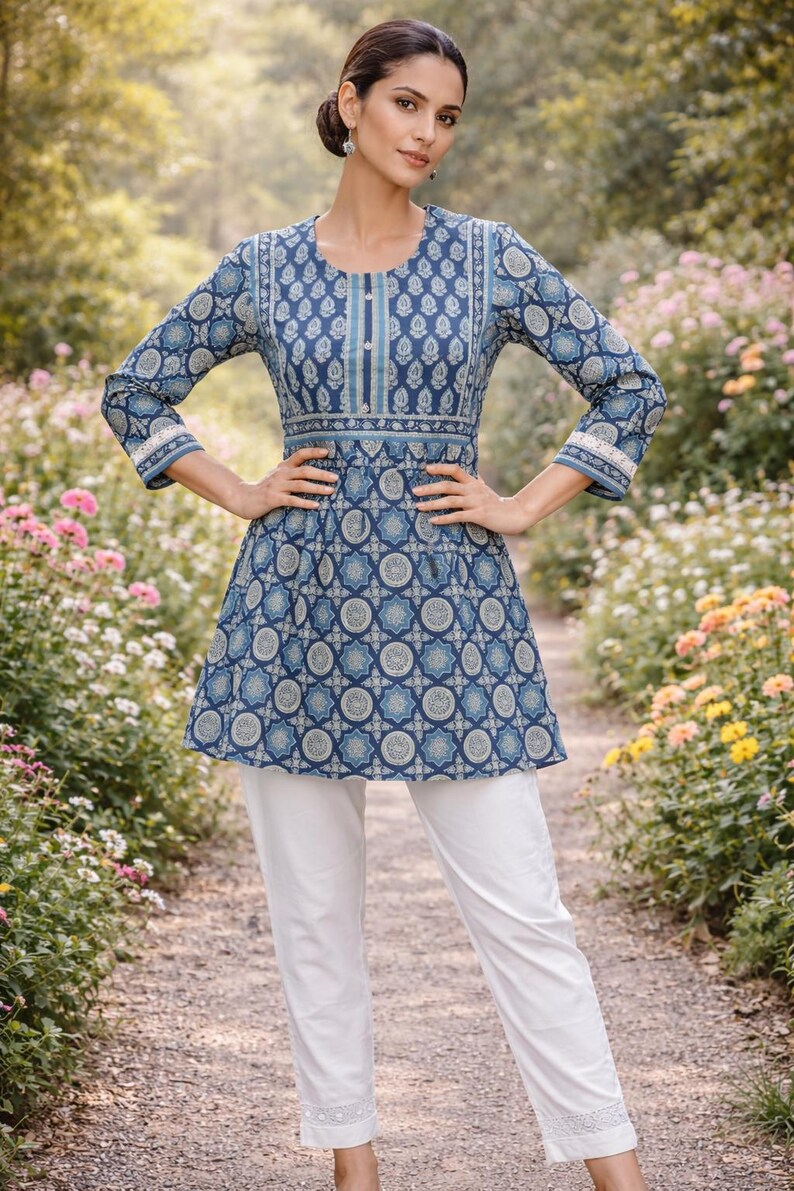 May include: A woman wearing a blue tunic with a geometric pattern and white pants. The tunic has long sleeves and a round neckline. The background is a garden with flowers.