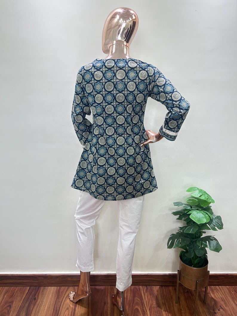 May include: A long-sleeved tunic and white pants ensemble. The tunic features a dark blue and light blue geometric pattern. The white pants have a decorative hem. The outfit is displayed on a mannequin.