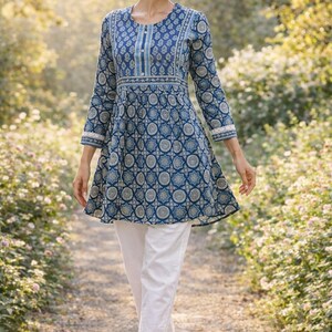 May include: A woman wearing a blue tunic with a white and blue circular pattern, paired with white pants. The tunic has long sleeves and a flared hem. The outfit is worn outdoors on a path with greenery in the background.