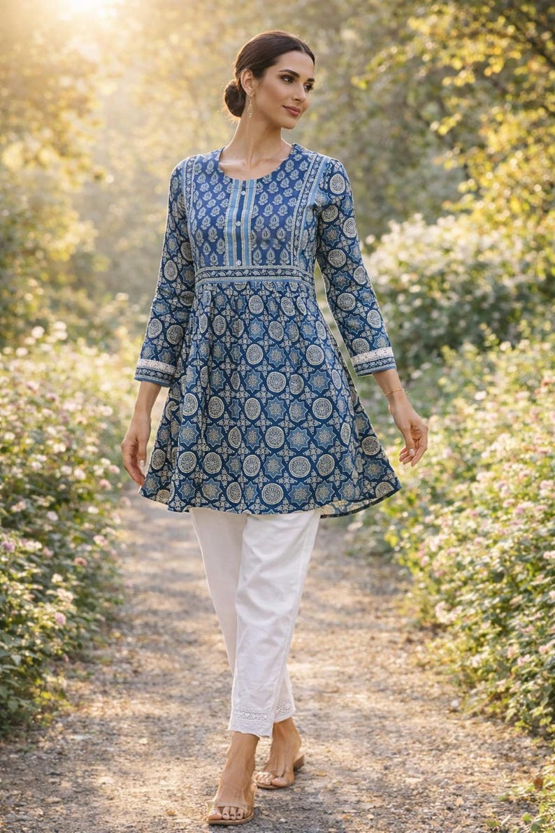 May include: A woman wearing a blue tunic with a white and blue circular pattern, paired with white pants. The tunic has long sleeves and a flared hem. The outfit is worn outdoors on a path with greenery in the background.