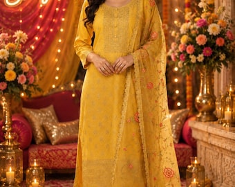 May include: A woman in a mustard yellow salwar kameez suit with a matching dupatta. The suit has detailed embroidery and a floral patterned dupatta. The word "Sangeet" is displayed in a neon sign.