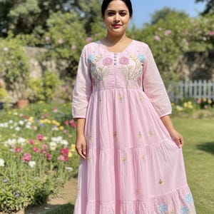 May include: A pale pink maxi dress with floral embroidery. The dress has a V-neck, three-quarter sleeves, and a tiered skirt. The embroidery features pink, blue, and yellow flowers. The dress is worn outdoors in a garden setting.
