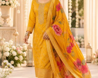 May include: A mustard yellow salwar kameez set featuring a long-sleeved tunic with intricate gold embroidery around the neckline and hem, matching straight-leg trousers, and a coordinating yellow dupatta with a vibrant pink and green floral print.