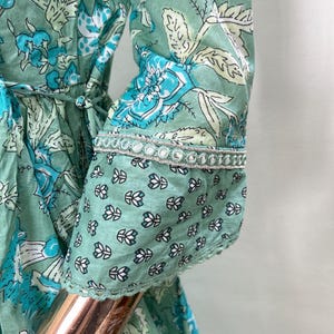 May include: A mint green dress with a floral pattern in shades of blue and white. The dress features long sleeves with a decorative trim at the cuffs. The dress has a tie at the waist and a tassel detail.