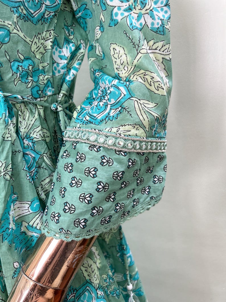 May include: A mint green dress with a floral pattern in shades of blue and white. The dress features long sleeves with a decorative trim at the cuffs. The dress has a tie at the waist and a tassel detail.