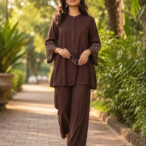 Könnte beinhalten: Ein braunes zweiteiliges Outfit, bestehend aus einer langärmligen Tunika und einer Hose mit weitem Bein. Die Tunika hat einen Stehkragen und gestickte Details an den Ärmeln. Das Outfit wird mit Sandalen und einer Brille getragen.