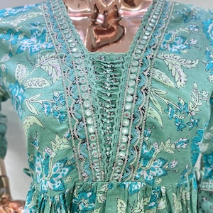 May include: A light green dress with a floral pattern in shades of blue and white. The dress features a V-neckline with intricate mirror and lace detailing. The sleeves are long and the dress has a gathered waist.