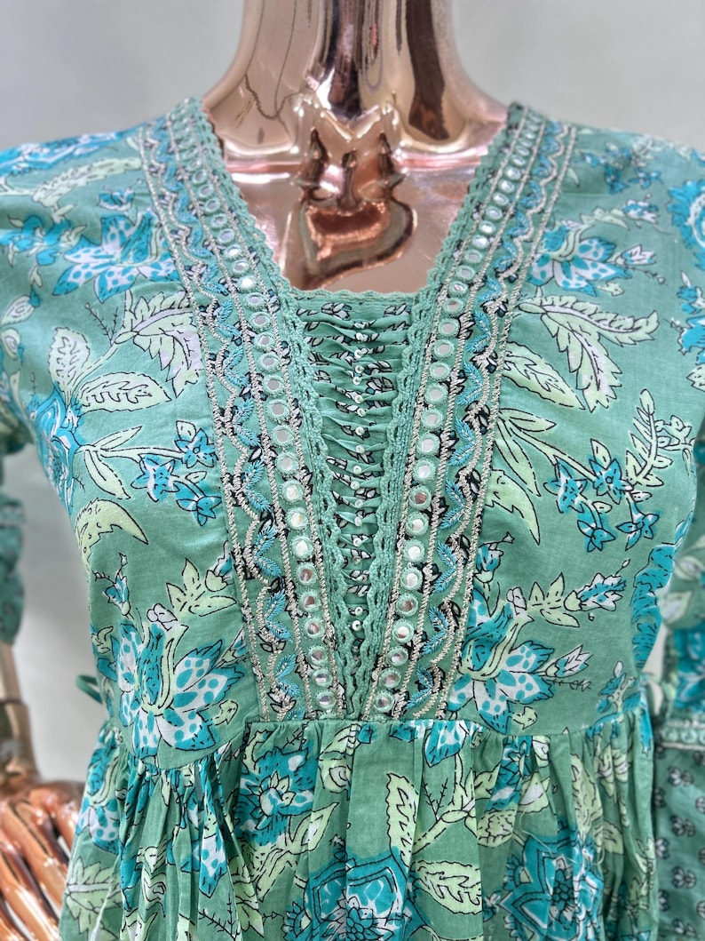 May include: A light green dress with a floral pattern in shades of blue and white. The dress features a V-neckline with intricate mirror and lace detailing. The sleeves are long and the dress has a gathered waist.