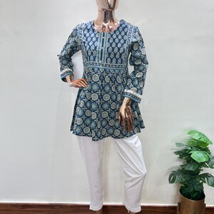 May include: A two-piece outfit on a mannequin. The top is a teal and white patterned tunic with long sleeves and a flared hem. The pants are white, cropped, and embroidered at the ankles. The outfit is displayed on a mannequin with a gold-colored head and hands.