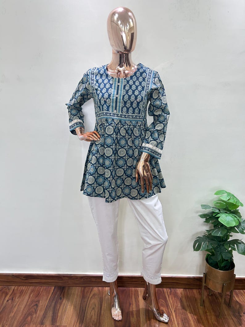 May include: A two-piece outfit on a mannequin. The top is a teal and white patterned tunic with long sleeves and a flared hem. The pants are white, cropped, and embroidered at the ankles. The outfit is displayed on a mannequin with a gold-colored head and hands.
