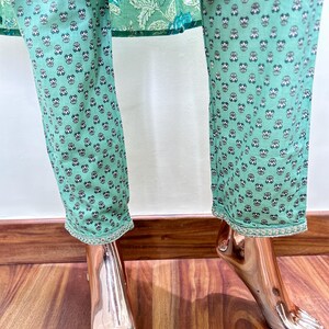 May include: Mint green pants with a repeating floral pattern in black and white. The pants have a decorative trim at the hem. The top of the pants is visible, featuring a floral design in shades of green and blue.