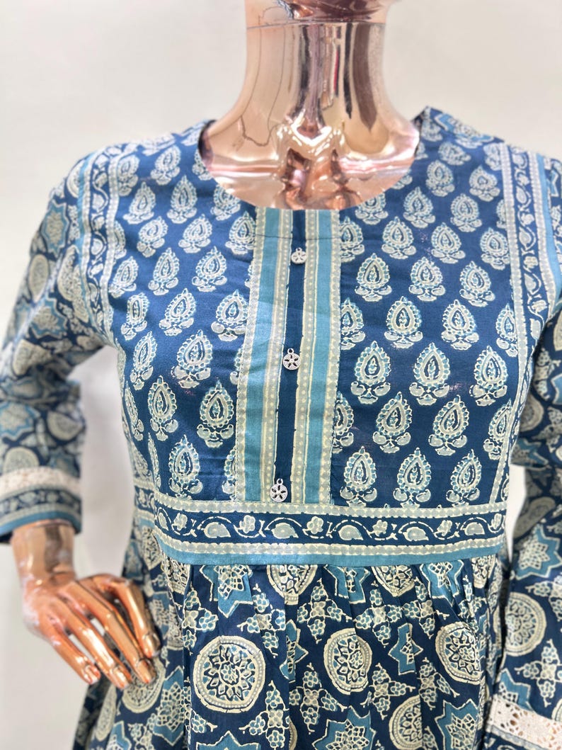 May include: A blue long-sleeve dress with a white paisley pattern. The dress features a round neckline, decorative buttons, and a contrasting stripe down the center. The sleeves have a lace trim.