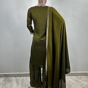May include: A dark green three-piece outfit with a long-sleeved top, palazzo pants, and a matching dupatta. The outfit has gold trim and is made of a soft, flowing fabric.