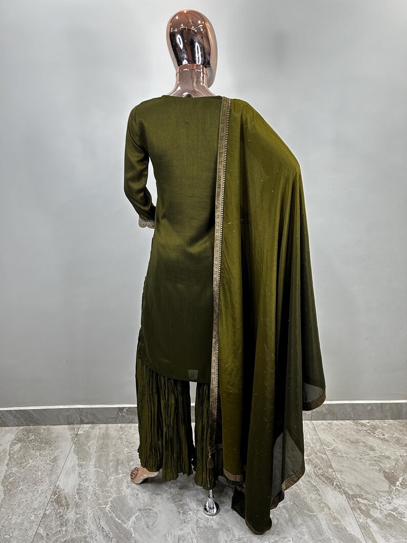 May include: A dark green three-piece outfit with a long-sleeved top, palazzo pants, and a matching dupatta. The outfit has gold trim and is made of a soft, flowing fabric.