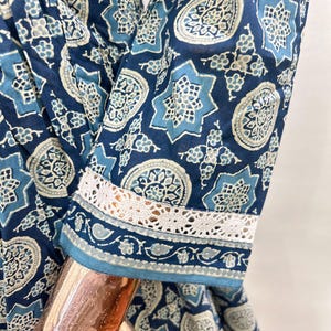 May include: Close-up of a blue patterned dress sleeve. The fabric features a repeating design of star and floral motifs in shades of blue and white. The sleeve cuff is trimmed with white lace.