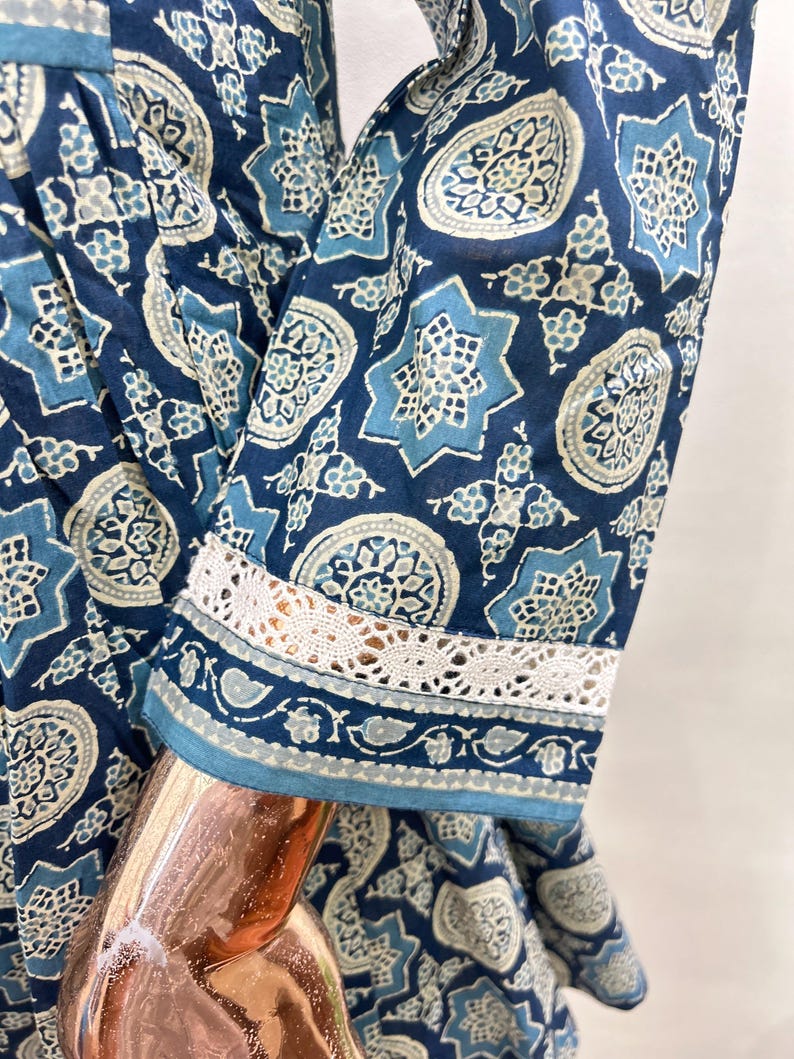 May include: Close-up of a blue patterned dress sleeve. The fabric features a repeating design of star and floral motifs in shades of blue and white. The sleeve cuff is trimmed with white lace.
