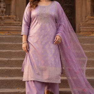 May include: A lilac-colored salwar kameez set. The outfit includes a long tunic with intricate embroidery around the neckline, matching wide-leg pants, and a sheer dupatta. The fabric has a subtle, textured pattern. The model is wearing light-colored heels.