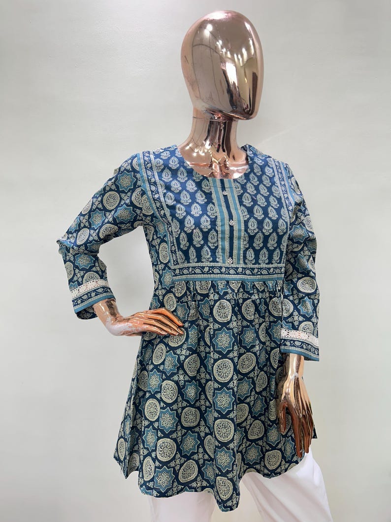 May include: A blue tunic with a white and blue pattern. The tunic has three-quarter sleeves with lace trim and a round neckline. The garment is displayed on a mannequin.