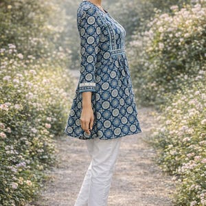 May include: A woman wearing a blue patterned tunic with three-quarter sleeves and white pants. The tunic features a V-neck and a flared hem. The pants are cropped and have embroidered details at the hem. The woman is wearing heeled sandals.