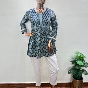 May include: A mannequin displays a two-piece outfit. The top is a dark teal tunic with a white and teal circular pattern, long sleeves, and white trim. The outfit is paired with white cropped pants. The mannequin is on a wooden floor.
