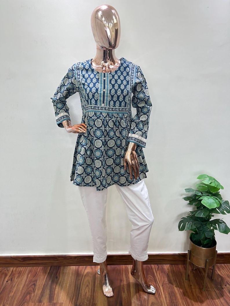 May include: A mannequin displays a two-piece outfit. The top is a dark teal tunic with a white and teal circular pattern, long sleeves, and white trim. The outfit is paired with white cropped pants. The mannequin is on a wooden floor.
