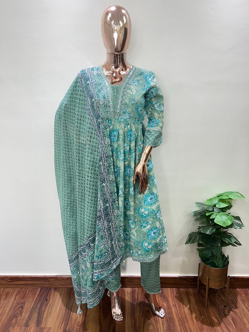 May include: A mint green floral print salwar kameez set displayed on a mannequin. The set includes a long-sleeved tunic, matching pants, and a patterned scarf. The tunic and pants feature a floral design in shades of blue and green.