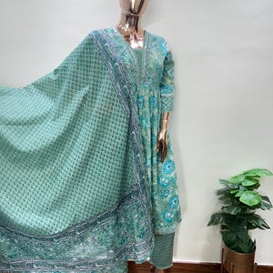 May include: A mint green Indian salwar kameez set displayed on a mannequin. The set includes a long tunic, matching pants, and a flowing scarf. The tunic and scarf feature floral patterns and decorative borders in shades of blue and green. The outfit is made of lightweight fabric.