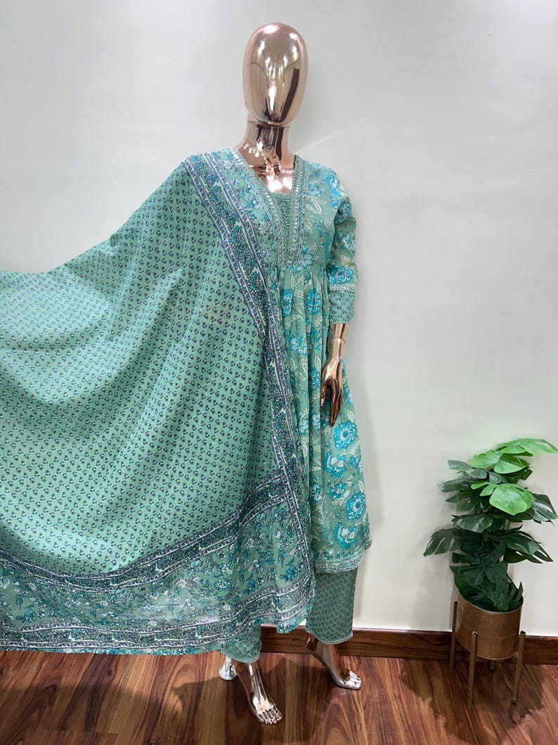 May include: A mint green Indian salwar kameez set displayed on a mannequin. The set includes a long tunic, matching pants, and a flowing scarf. The tunic and scarf feature floral patterns and decorative borders in shades of blue and green. The outfit is made of lightweight fabric.
