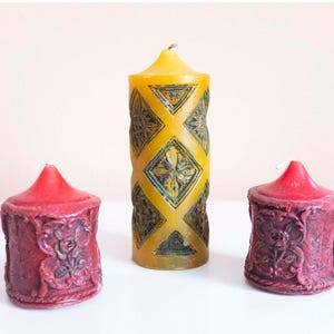 May include: Three decorative pillar candles. One tall yellow candle with a diamond pattern and two smaller red candles with floral designs. The candles are on a white surface.