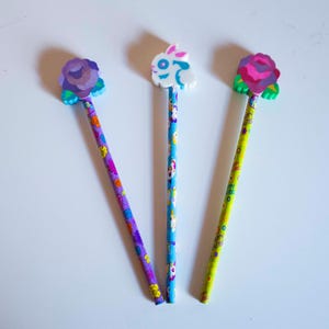May include: Three decorative pencils with eraser toppers. One pencil has a purple rose, another a white bunny, and the third a pink rose. The pencils have colorful floral patterns on the shafts.