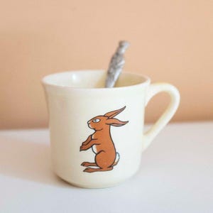 May include: Cream ceramic mug with a brown rabbit illustration. The mug has a curved handle and a silver spoon inside. The rabbit is depicted in a seated position with its ears perked up. A charming and whimsical design, perfect for a hot beverage.