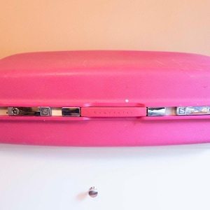 Puede incluir: Una maleta rígida de color rosa brillante con forma rectangular y esquinas redondeadas. El equipaje cuenta con un asa central, dos cierres metálicos plateados con cerraduras y un borde plateado a lo largo de la costura central.