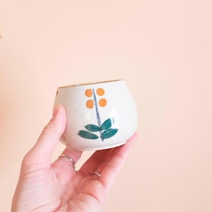 Może przedstawiać: Mały, kremowobiały ceramiczny kubek z kwiatowym wzorem. Wzór przedstawia pomarańczowe kółka i zielone liście na niebieskiej łodydze. Kubek trzymany jest w dłoni na brzoskwiniowym tle.