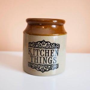May include: A ceramic kitchen jar with a brown lid and a beige body. The jar has the words "KITCHEN THINGS" in black lettering with decorative scrollwork. The jar is on a white surface.