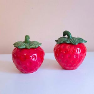 May include: Two ceramic strawberry-shaped containers with glossy red bodies and textured surfaces. Each piece features a green leafy stem lid on top. The containers are rounded and bulbous, mimicking the appearance of fresh strawberries.