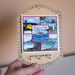 Puede incluir: Un porta llaves de madera con una baldosa que presenta una ilustración vintage de las Cataratas del Niágara y el texto "Niagara Falls". El porta llaves tiene cuatro ganchos y el texto "Watch Your Keys".