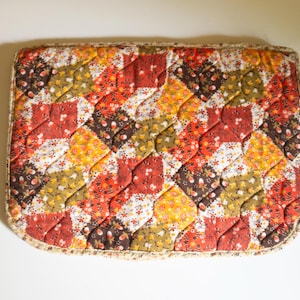 May include: A quilted patchwork cushion with a floral pattern in shades of red, orange, yellow, brown, and white. The cushion has a fringed border.
