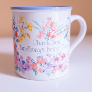 Puede incluir: Taza de cerámica blanca con borde y asa azul claro, adornada con un diseño floral en tonos rosa, amarillo y azul. La taza presenta el texto "Thank You For Always Being There" en una fuente cursiva.