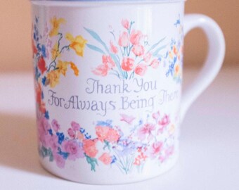 Könnte beinhalten: Weiße Keramiktasse mit hellblauem Rand und Henkel, verziert mit einem Blumenmuster in Rosa-, Gelb- und Blautönen. Die Tasse trägt den Schriftzug "Thank You For Always Being There" in Schreibschrift.