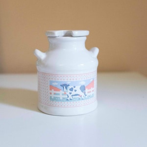 Puede incluir: Jarra de leche de cerámica blanca con tapa y dos asas pequeñas. La jarra presenta una banda decorativa con una vaca, una valla y una escena de granja. Una adición encantadora a una cocina de estilo rústico.