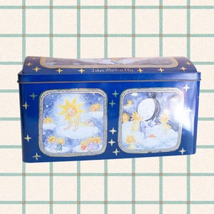 Könnte beinhalten: Eine blaue Metallbox mit Illustrationen von Sonne und Mond. Die Box hat eine gewölbte Oberseite und zeigt ein himmlisches Thema mit Sternen und dem Text "Ida Bohatta". Die Illustrationen zeigen ein Sonnenbaby und eine Mondfigur.