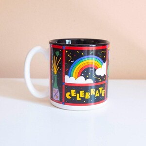 Può includere: Una tazza in ceramica con manico bianco e interno nero. L'esterno presenta uno sfondo nero con un arcobaleno colorato, nuvole bianche, coriandoli e l'illustrazione di una bottiglia di champagne. La parola CELEBRATE è stampata in lettere gialle.